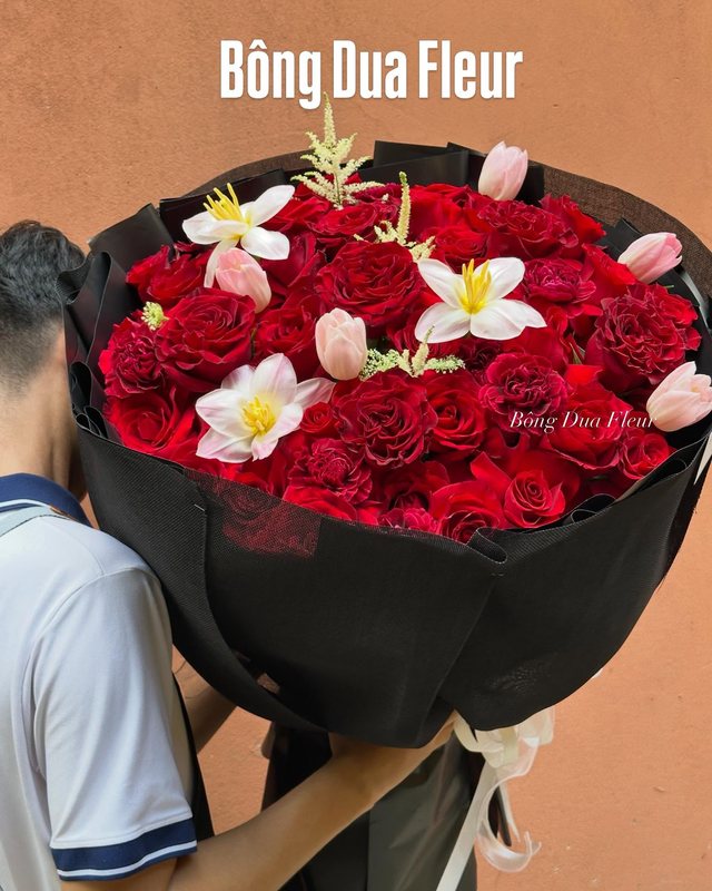 Bó hoa hồng đỏ điểm lily trắng, gói giấy đen, được một người ôm trước ngực.