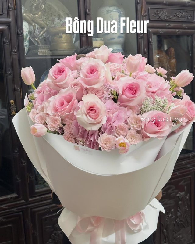 Bó hoa hồng phấn lớn gói giấy trắng, đặt trên nền tối.