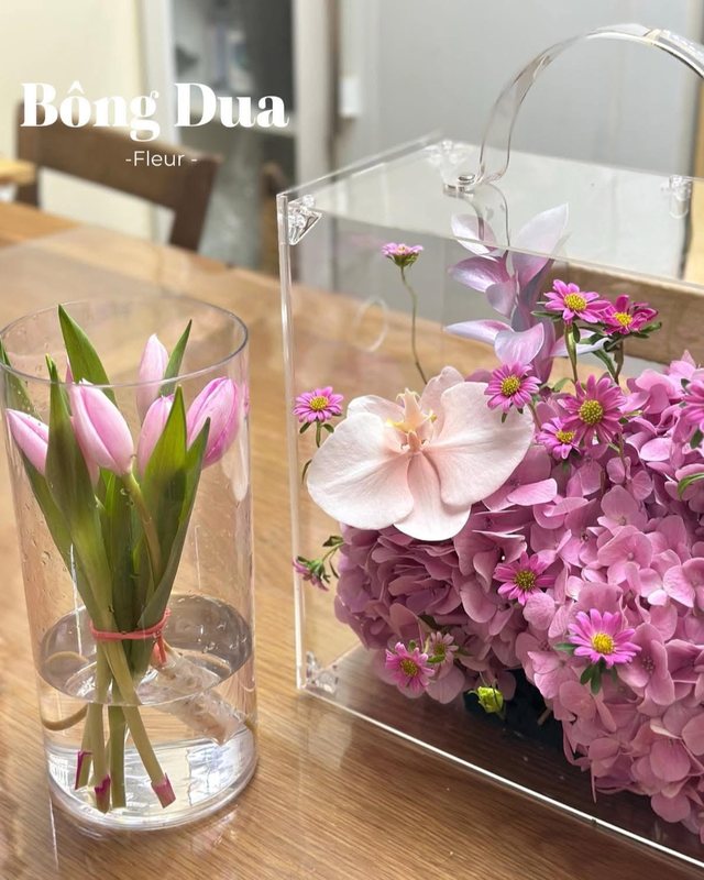 Bình thuỷ tinh cắm tulip hồng bên cạnh hộp mica trong suốt cắm hoa lan tím.