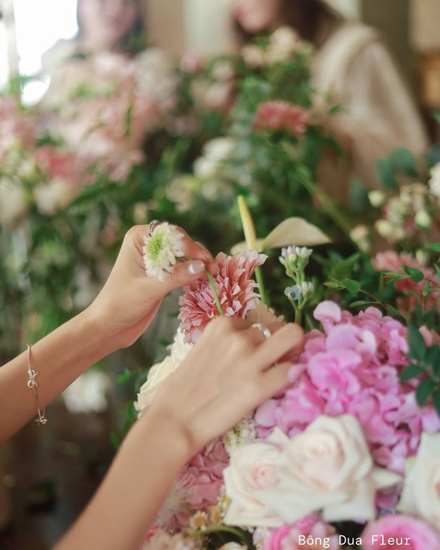 Đôi bàn tay đang chạm vào một cành hoa trắng giữa bàn hoa tươi.